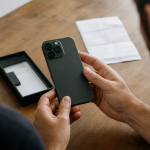 Pessoa conferindo o IMEI de um celular usado antes da compra, com aparelho e nota fiscal sobre uma mesa, sem textos legíveis.