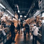 Corredor de mercado interno movimentado, com lojas, pessoas circulando e consumidores fazendo compras.