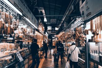 Corredor de mercado interno movimentado, com lojas, pessoas circulando e consumidores fazendo compras.