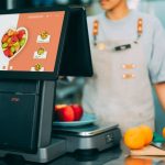 Sistema digital de checkout em uso no caixa de um supermercado, com frutas ao redor e uma caixa ao fundo.