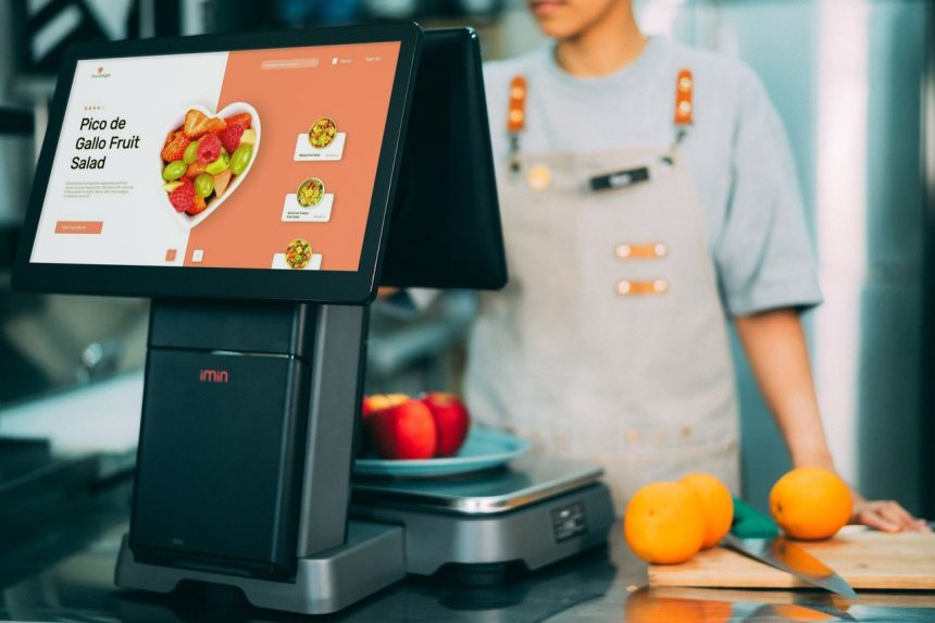 Sistema digital de checkout em uso no caixa de um supermercado, com frutas ao redor e uma caixa ao fundo.